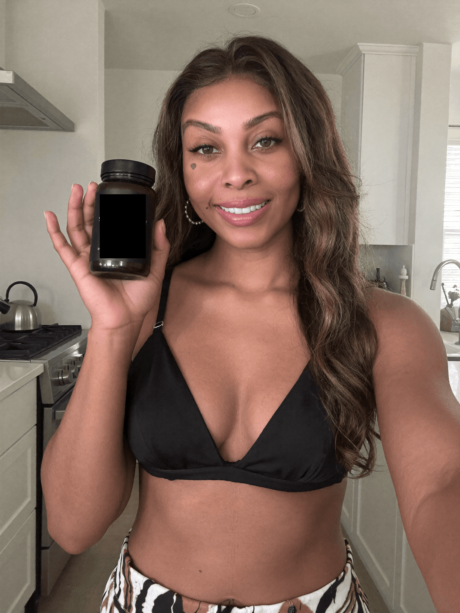 Smiling woman with long brown hair holds a black bottle in a bright, modern kitchen.
