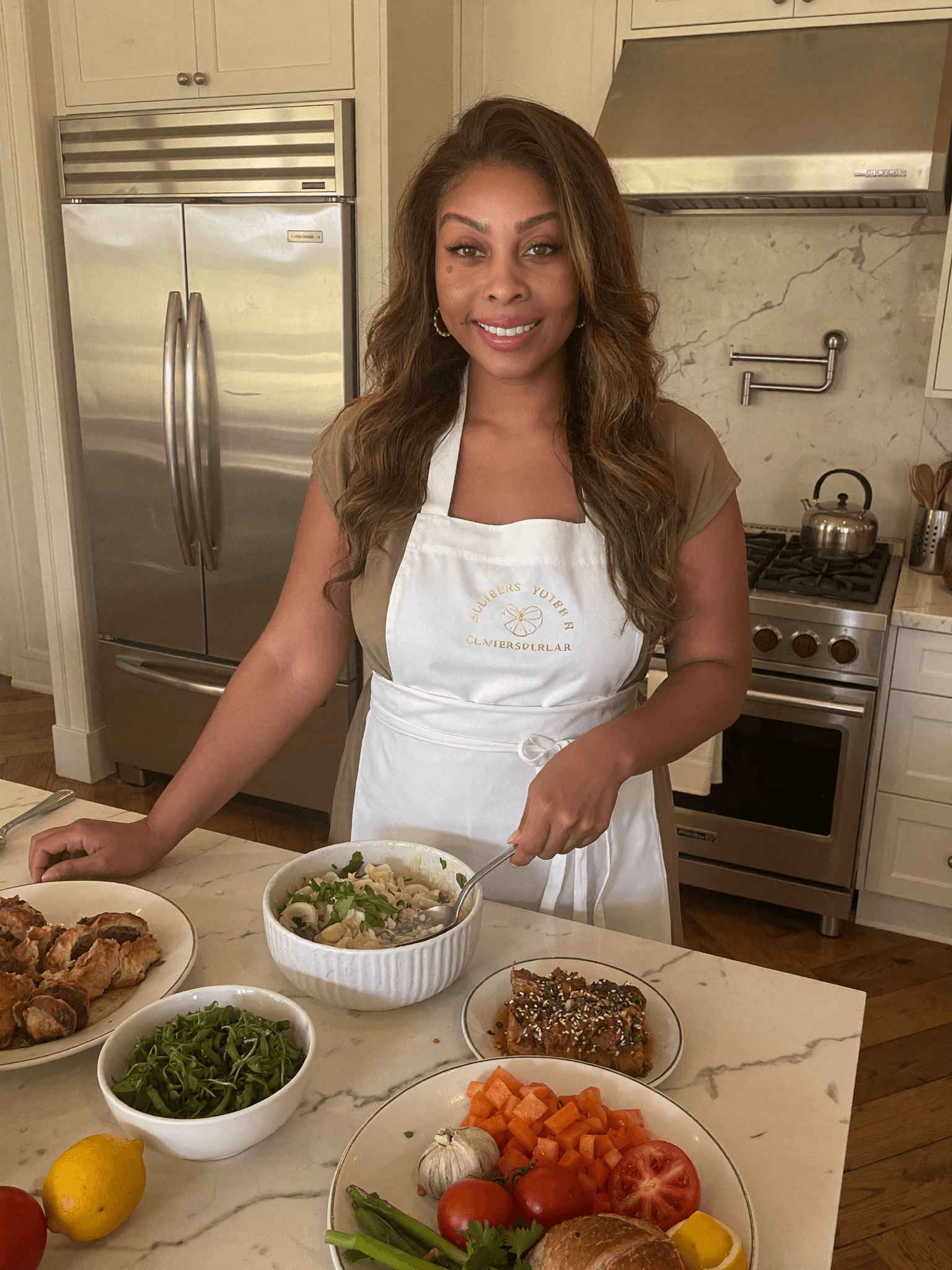 Smiling woman in a white apron prepares various dishes on a marble kitchen counter.