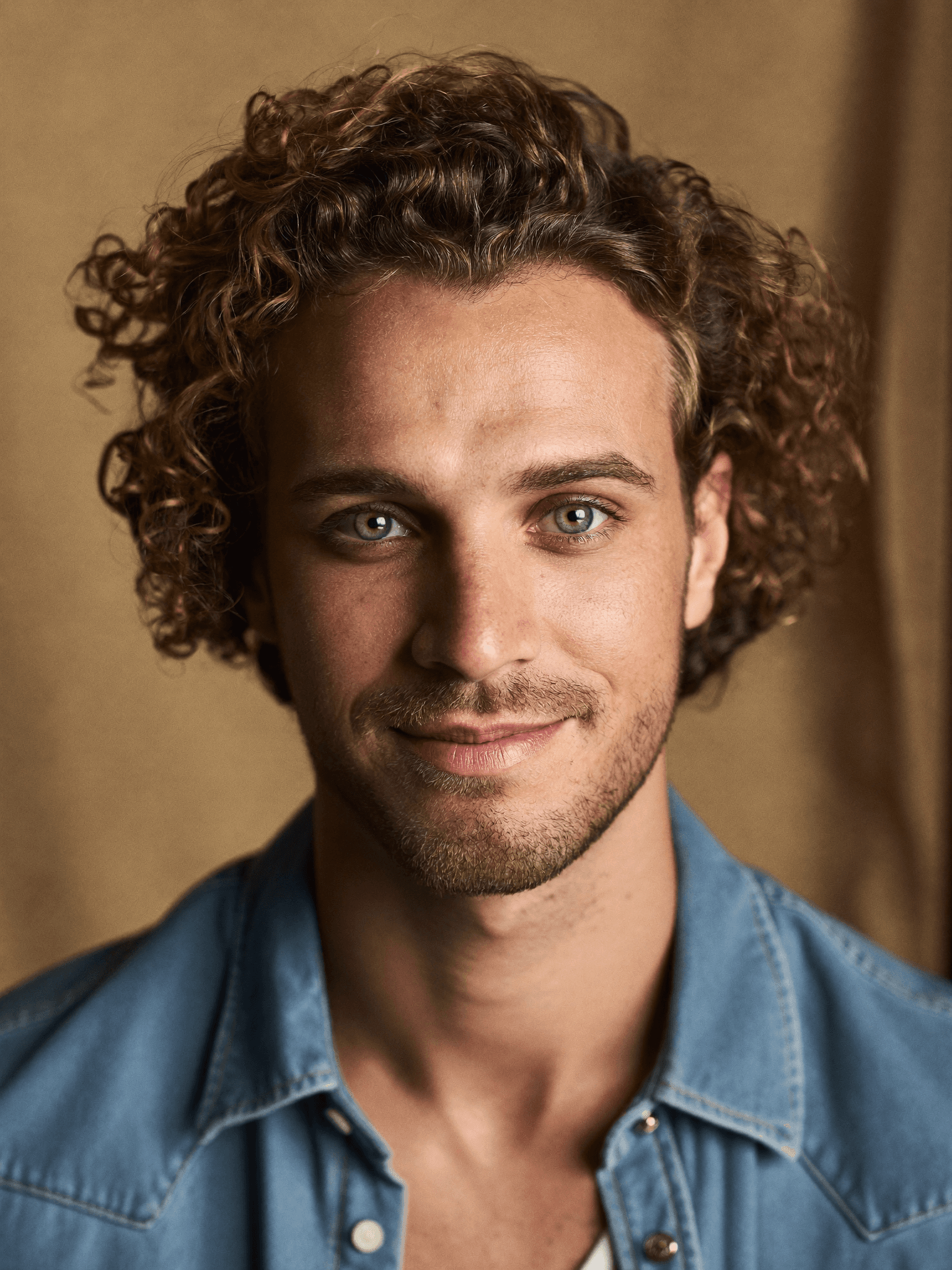 Smiling man with curly brown hair and striking green eyes wearing a blue denim shirt.