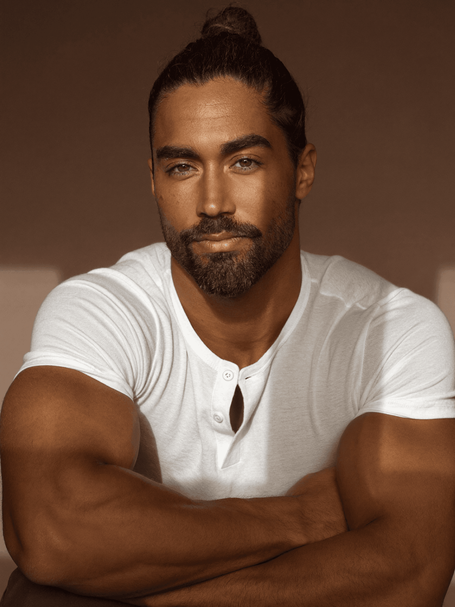 Muscular man with a beard and man bun wearing a white henley, arms crossed.