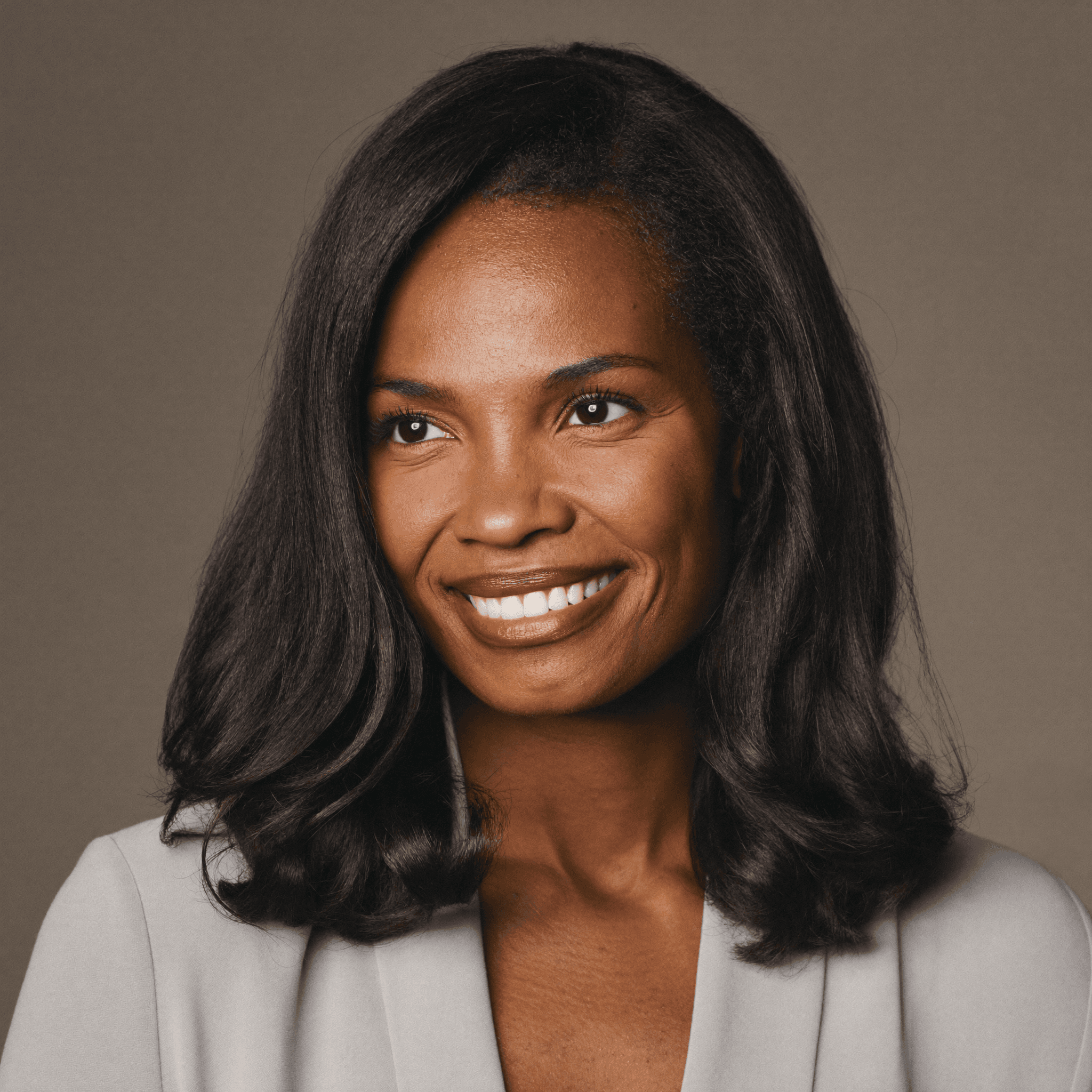Smiling Black woman with shoulder-length dark hair wearing a grey blazer against a brown background.