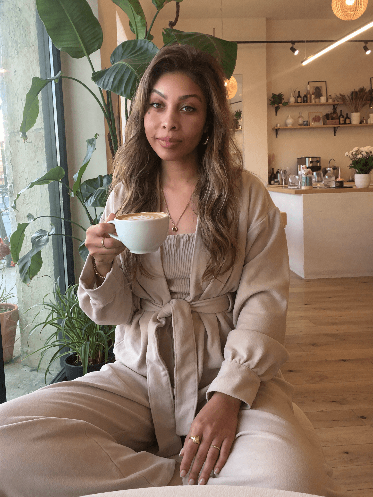 Woman in a beige lounge set holds a latte in a bright, plant-filled cafe.